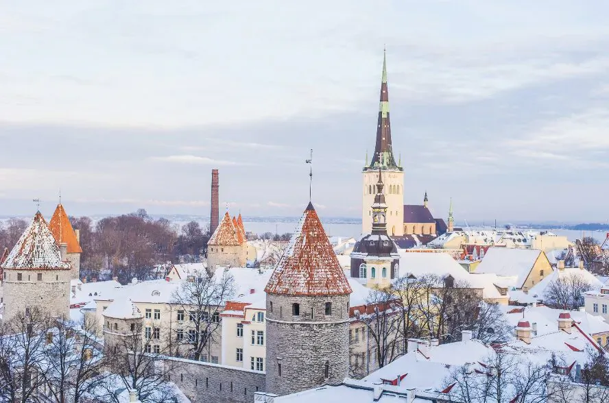 Winter panorama of Tallinn's Old Town with snow-covered red-roofed towers and tall green-spired church.