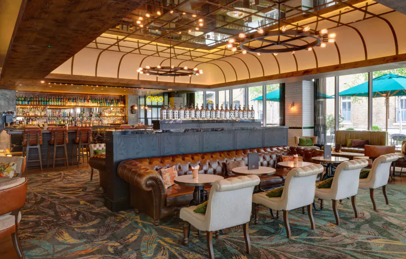 Elegant Gallery bar at Hilton London Bankside with ornate ceiling, leather sofas, round tables, bar, and green patio views.