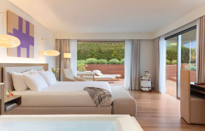 Luxurious hotel suite with king bed, jacuzzi, lounge chair on terrace overlooking lush green garden.