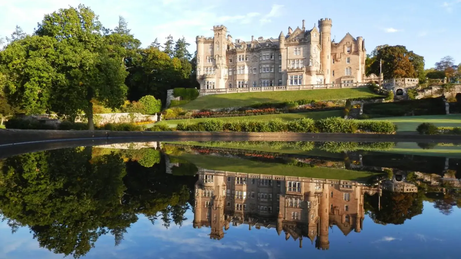 Grand castle with towers reflected perfectly in calm lake, surrounded by green lawns and trees at sunset.