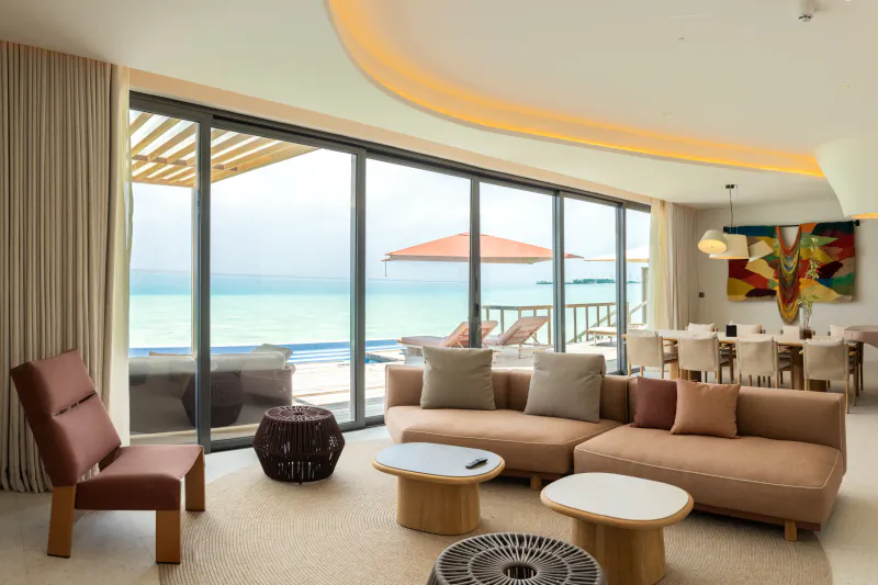 Luxurious SO/ Maldives villa living room with curved ceiling, plush sofas, dining table, and panoramic ocean views through glass doors.