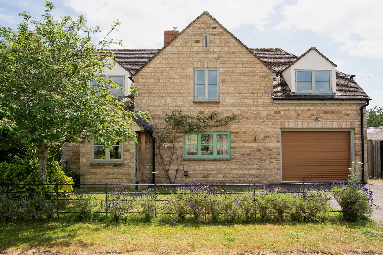 Timeless Cotswolds country house in Kingham with stone facade, garage, trees, and garden under partly cloudy sky
