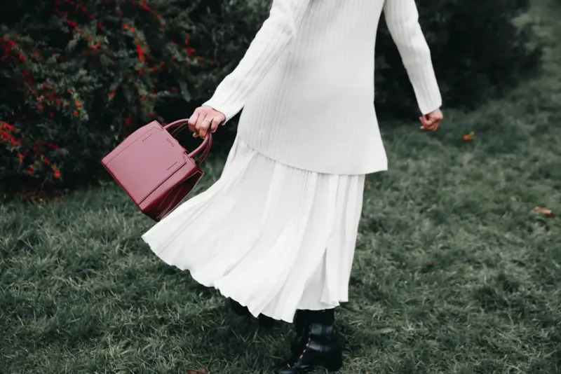Woman in white flowing dress and boots walks in grass, holding red handbag amid red-berried bushes