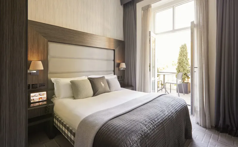 Luxury hotel bedroom with king bed, neutral linens, bedside lamps, and balcony doors open to garden terrace.