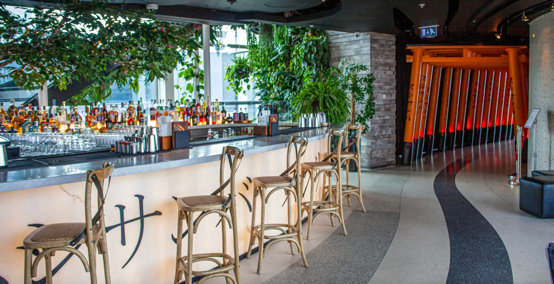 SushiSamba bar with wooden stools, lit bottles, plants, large windows, and orange torii gate backdrop