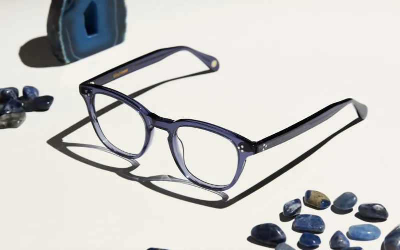 Navy blue Bloobloom eyeglasses displayed on white surface with scattered blue agate stones and large geode.