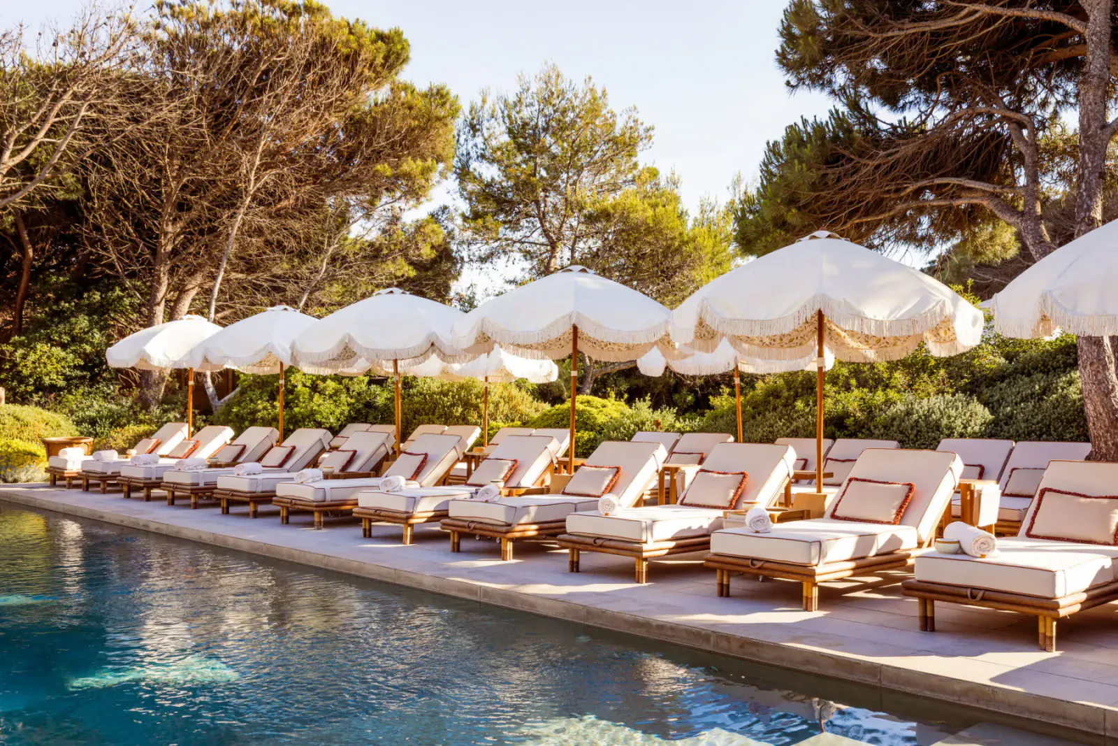 Row of white umbrellas shading wooden lounge chairs beside a luxurious pool at Gigi Rigolatto beach house, surrounded by pine trees.