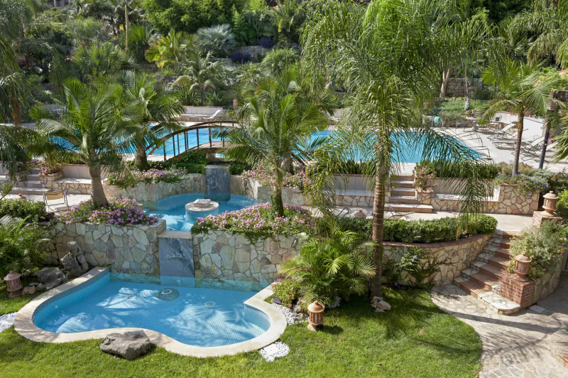 Aerial view of Wish Sicily luxury villas with turquoise pools, arched bridge, waterfalls, and lush palm gardens.