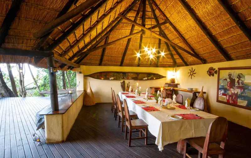 Elegant thatched dining area in Botswana safari lodge with long white-clothed table set for dinner, ocean view deck, and African art.