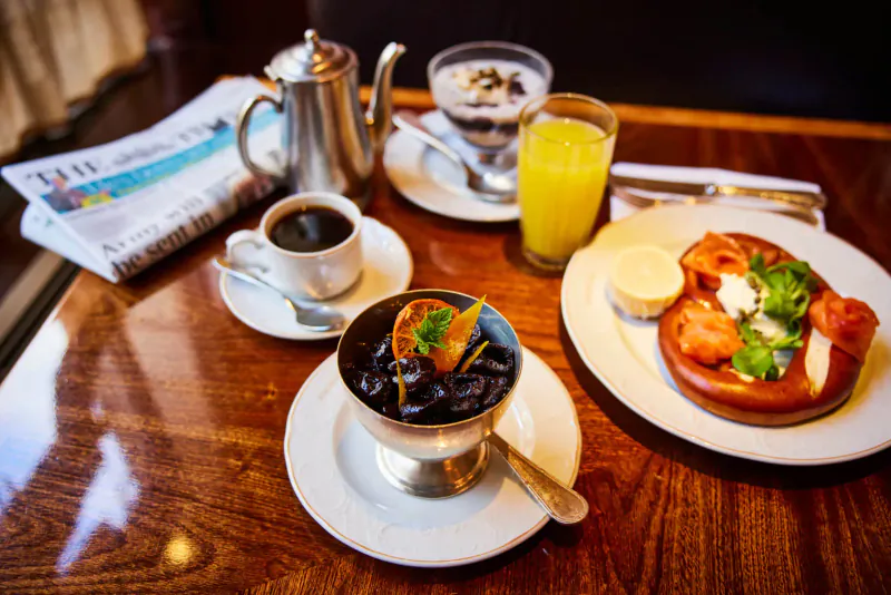 Elegant breakfast table: newspaper, coffee, tea pot, yogurt, orange juice, black pudding parfait, and pretzel with cream.