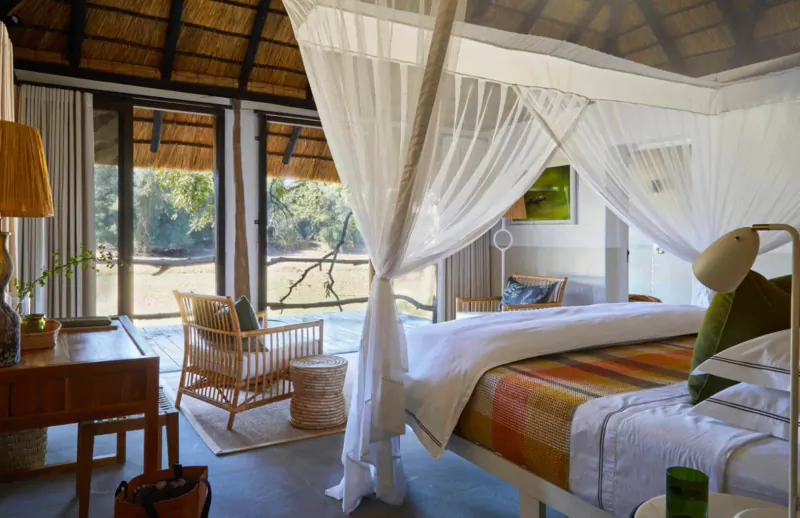 Luxurious thatched safari lodge bedroom with canopy bed, orange quilt, chairs, and bush view through open doors.