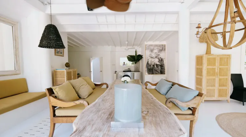 Bright Bali villa living room with yellow cushioned benches, rustic wood dining table, blue vase, rattan lamps, and plants.