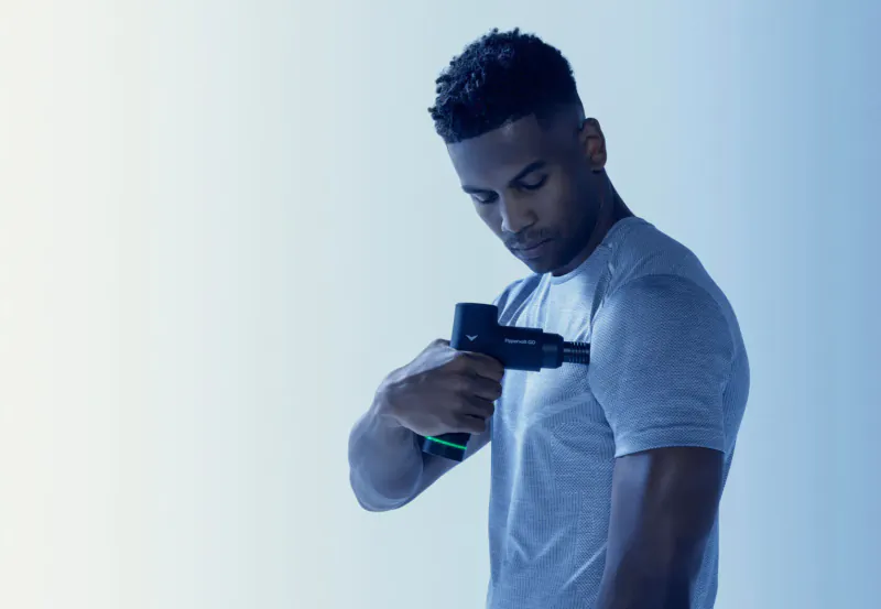 Black man in gray shirt using black Hypervolt GO massage gun on his shoulder against blue background