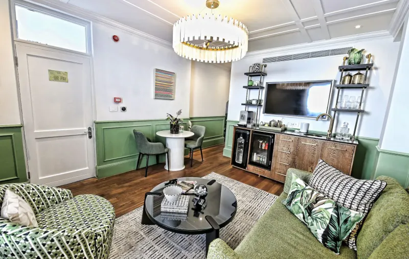 Elegant lounge with green armchairs, round table, TV, chandelier, and plants in modern room.