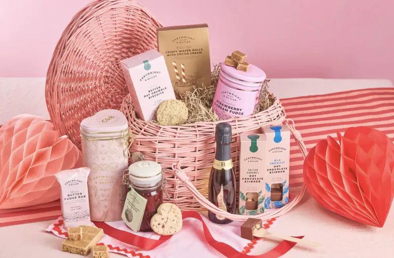 Open pink woven luxury gift basket with champagne, shortbread, fudge, chocolates, and pink paper fans on pink backdrop.