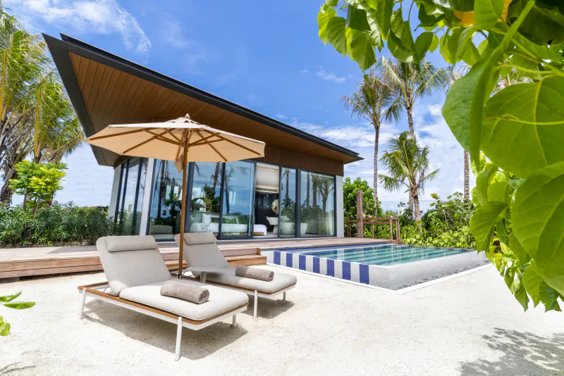 Modern beachfront villa with infinity pool, lounge chairs, umbrella at Crossroads Maldives, surrounded by palms and ocean.