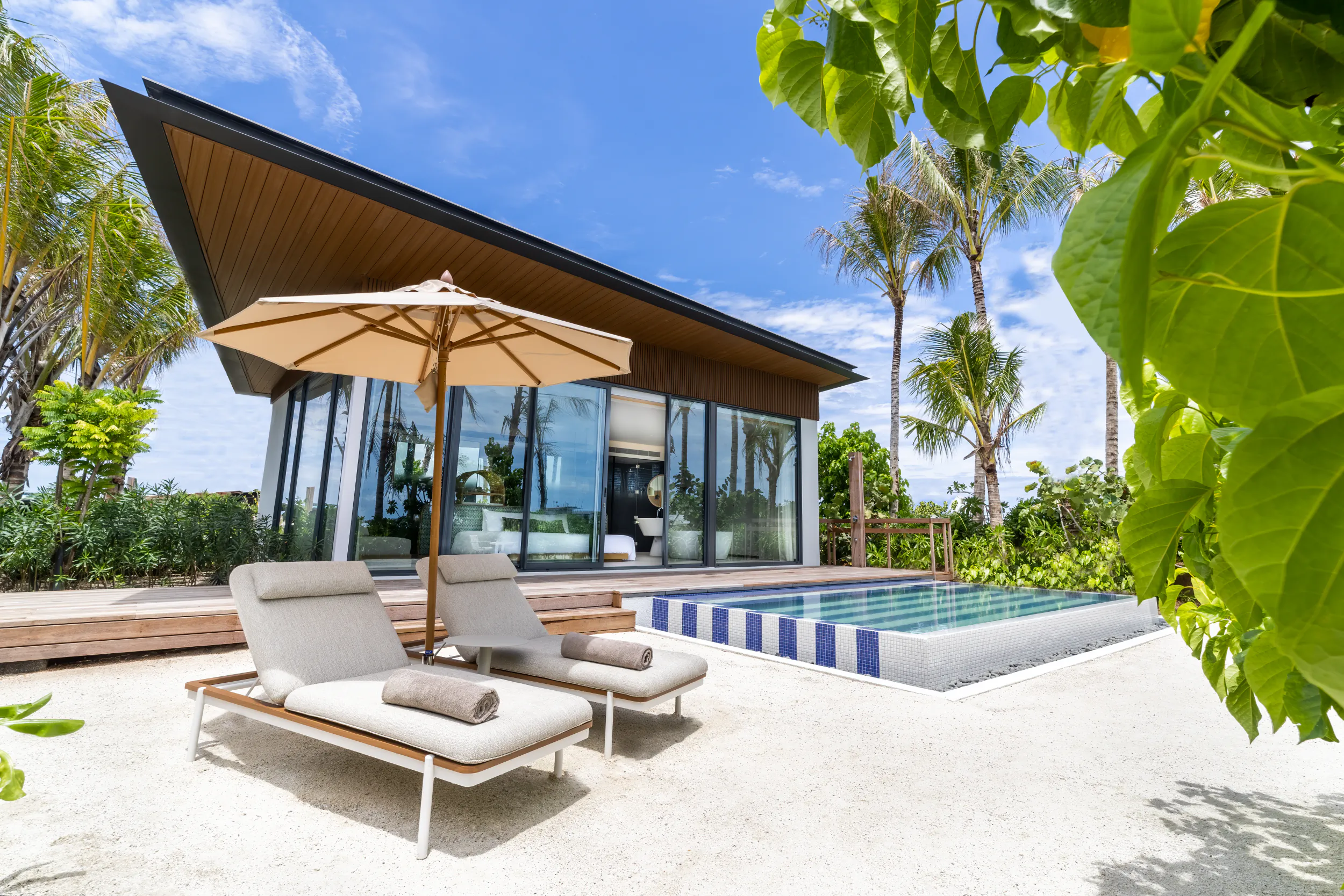 Modern beachfront villa with infinity pool, lounge chairs, umbrella at Crossroads Maldives, surrounded by palms and ocean.