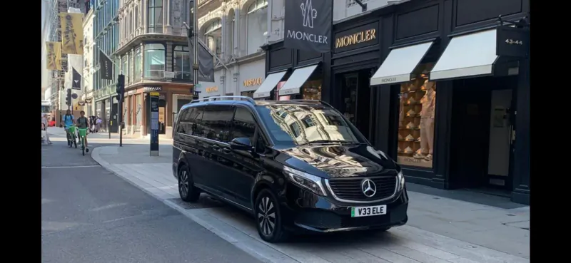 Black Mercedes V-Class van parked on city street outside Moncler luxury store, pedestrians nearby.