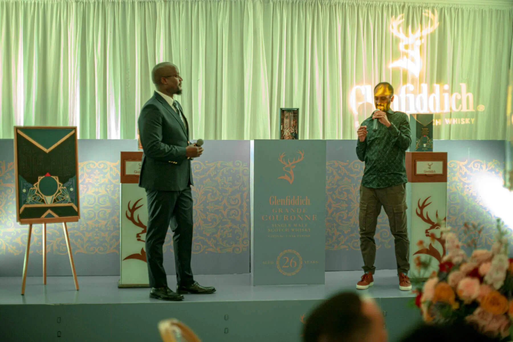 Two men on stage at Glenfiddich event: one in suit, one in green jacket, holding mics, deer logo backdrop.