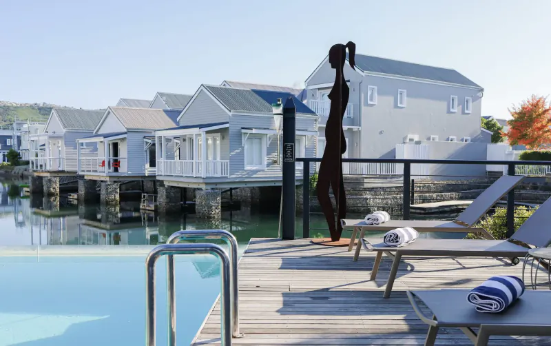 Silhouette of woman by infinity pool on wooden deck with lounge chairs, overlooking white waterfront houses at Turbine Hotel.