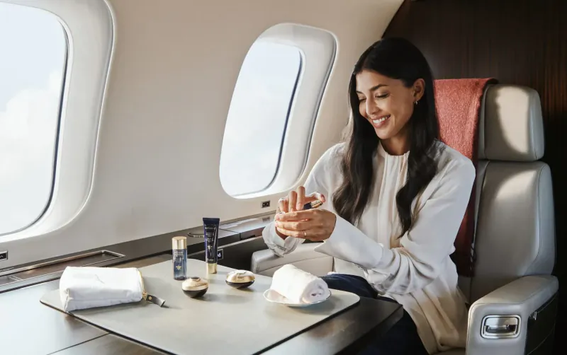 Smiling woman in white sweater applies skincare products at table in luxury airplane cabin with window view.