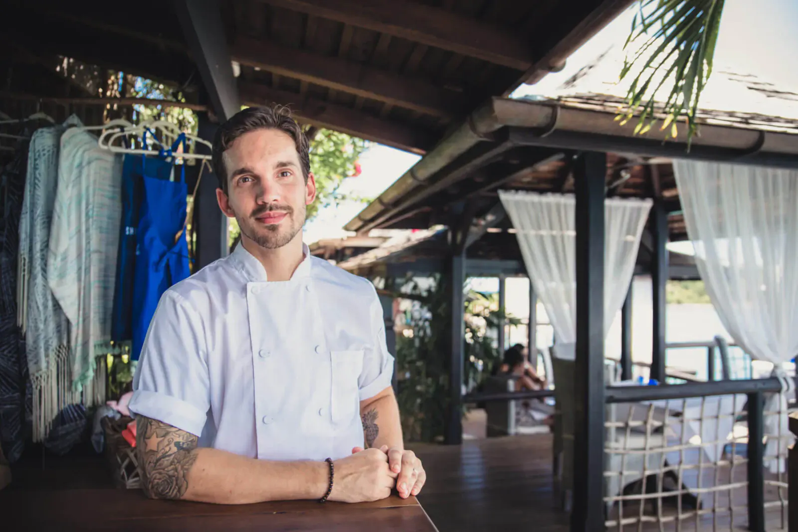 Executive Chef Simon Christey-French in white chef's jacket, tattooed arms, smiling at tropical deck restaurant with hanging clothes and sheer curtains.