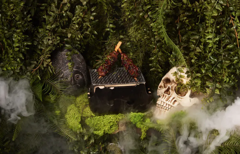 Halloween display: black box with red skewer, skull, stone head on mossy base amid ferns and mist in Wonderland style