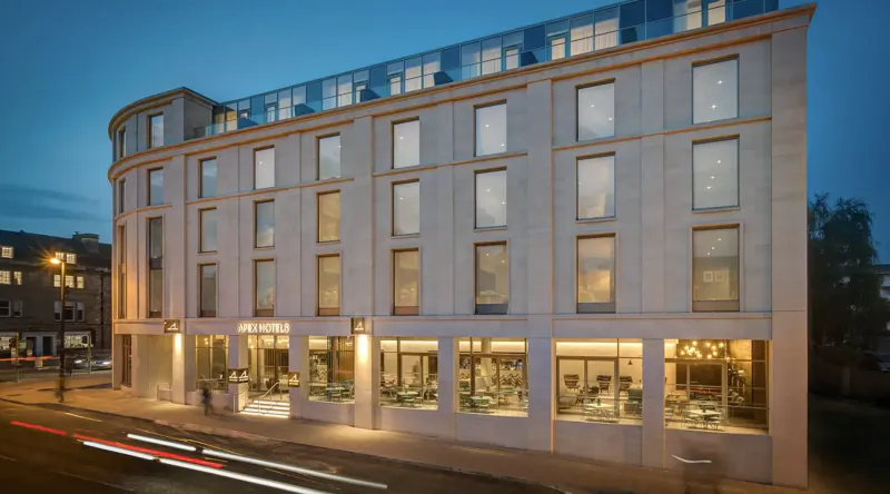 Apex City of Bath Hotel, modern multi-story building with glass facade at dusk in city center, light trails on street.