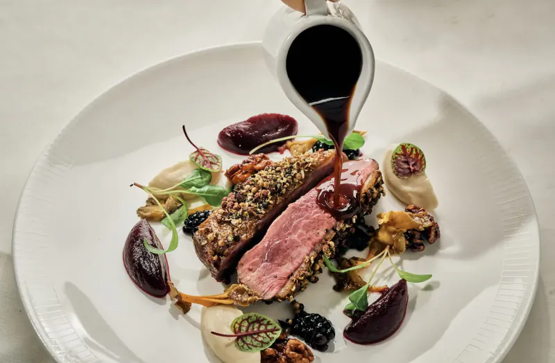 Close-up of sliced pink duck breast on white plate with blackberries, beets, nuts, greens, and sauce being poured from gravy boat.