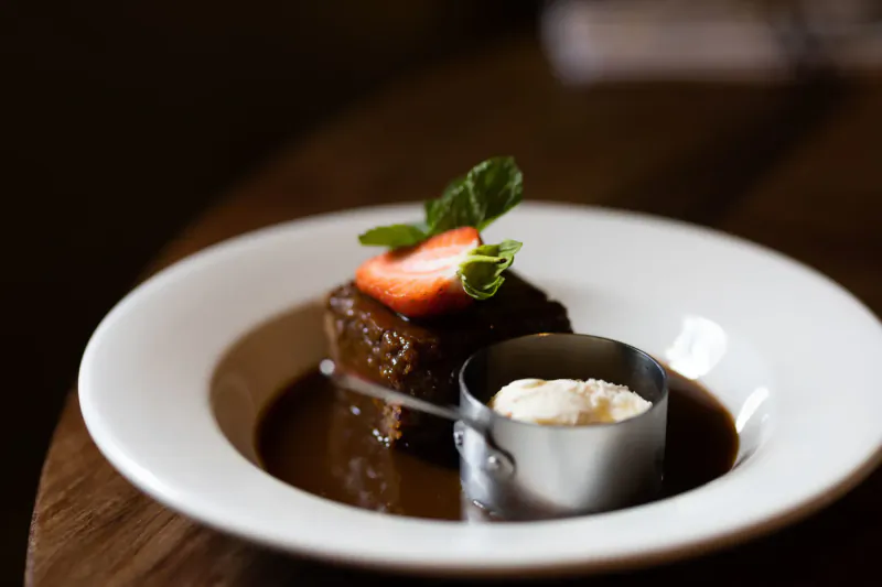 Sticky toffee pudding on plate with toffee sauce, strawberry, mint sprig, and side of cream on wooden table