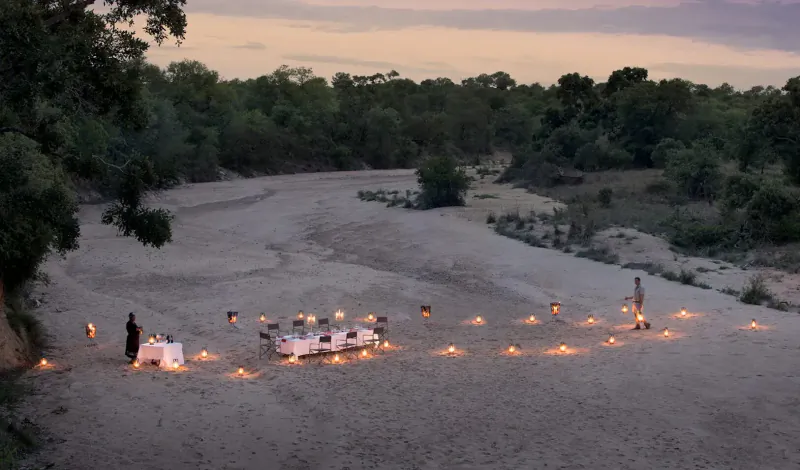 Romantic candlelit dinner table on sandy riverbank at sunset, surrounded by bushveld trees, two people standing nearby.