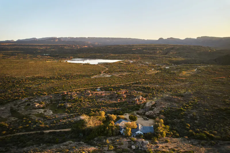 Aerial view of Bushman’s Kloof: blue-roofed lodge amid golden grasslands, lake, and rugged mountains at sunset.