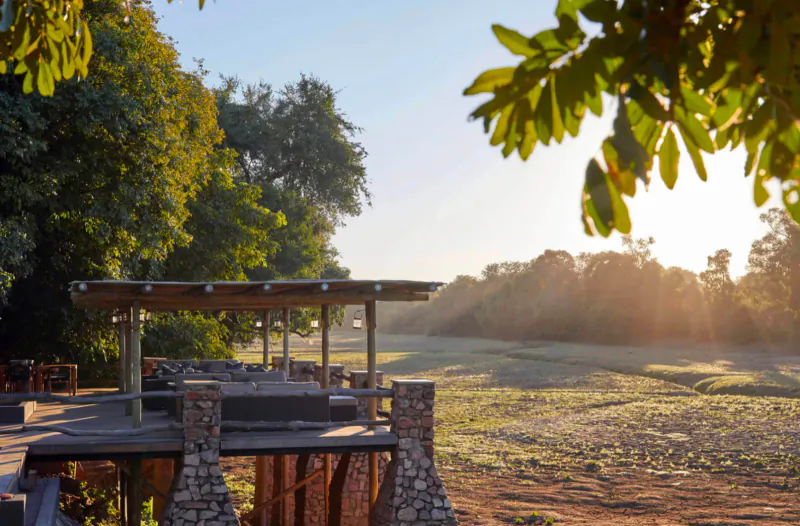 Wooden deck with lounge seating at Mfuwe Lodge, overlooking grassy savanna at sunset, framed by green trees.