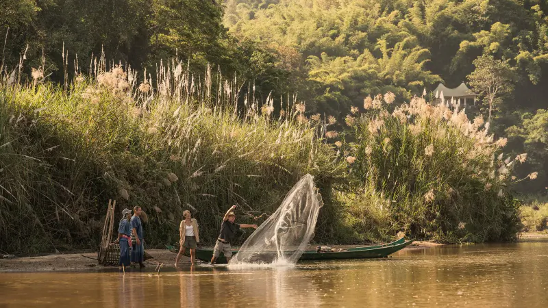 Three people casting a net from a green canoe on a lush riverside, surrounded by bamboo and reeds, Summer Family Fun with Four Seasons Thailand.