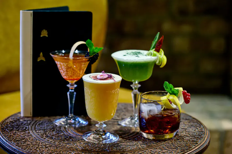 Four colorful cocktails garnished with mint, lemon, and chili on ornate tray beside black menu book at Indian restaurant.