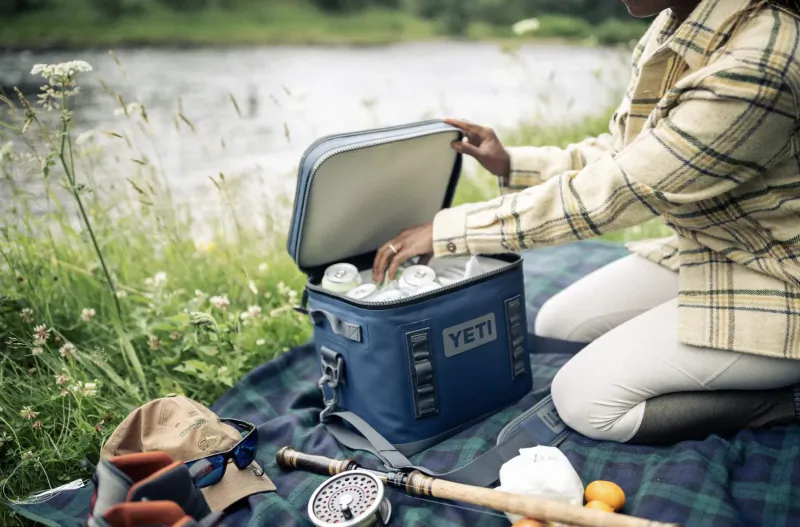 Woman opening blue YETI cooler with ice on plaid blanket by river, fishing rod nearby