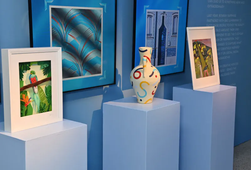 Gallery display: parrot painting, abstract blue art, numbered vase on pedestal, and landscape painting at AI art event.