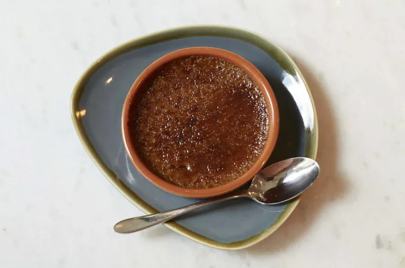 Crème brûlée in terracotta ramekin with silver spoon on teal plate, white surface.