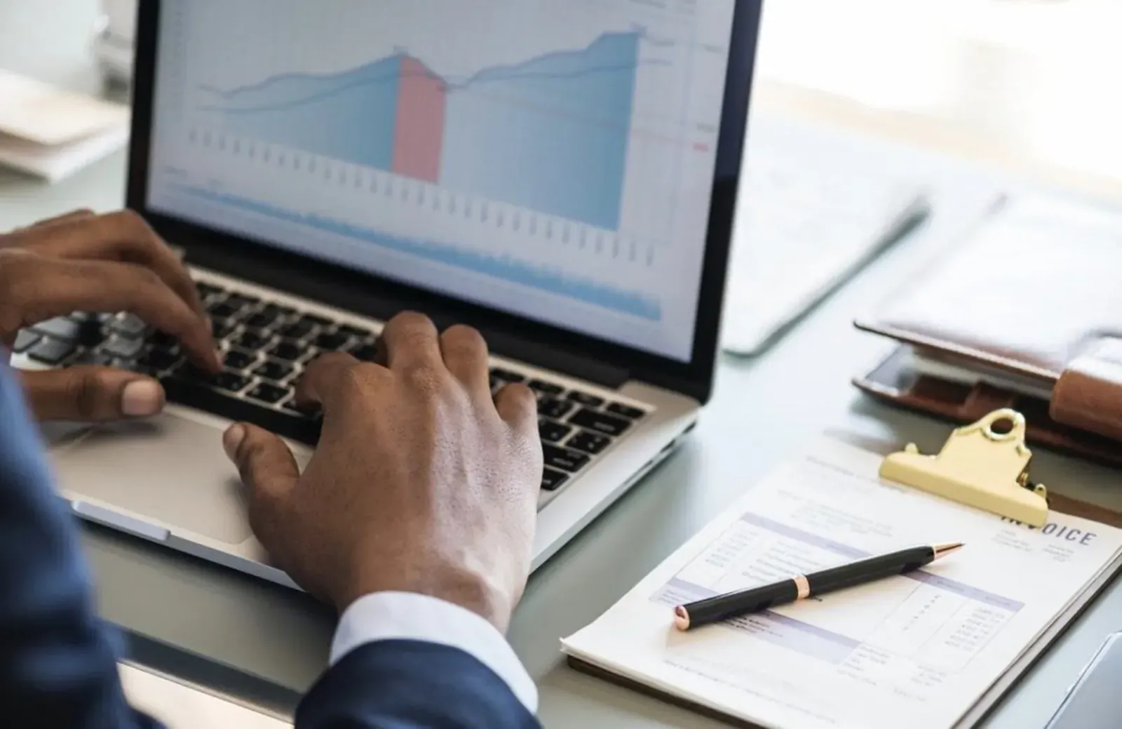 Close-up of Black man's hands typing on MacBook displaying rising financial charts, with notepad and pen nearby