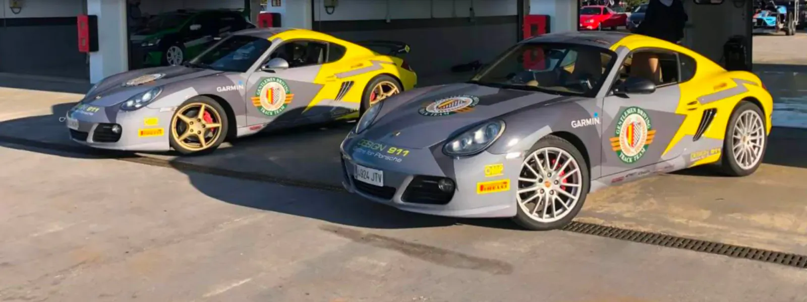 Two gray Porsche sports cars with yellow accents and Gentlemen Driving Club logos parked in a garage.