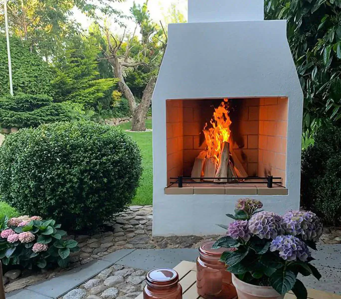 Schiedel Isokern Volcanic white brick garden fireplace with burning logs, surrounded by lush greenery, hydrangeas, and patio table.