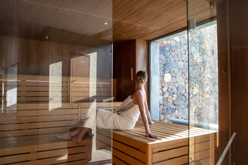 Woman wrapped in white towel sits on wooden bench in modern sauna with stone wall view, Jumeirah Port Soller Hotel & Spa.