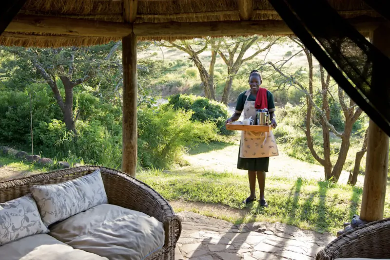 Woman in colorful headwrap and apron holding tray with bottles in thatched open-air lodge, lush Kenyan savanna backdrop