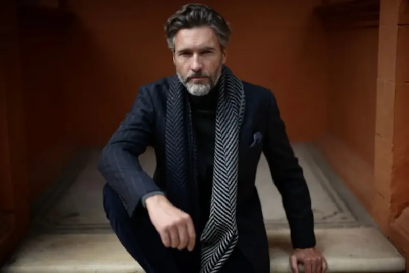 Silver-haired man in navy bespoke suit and patterned scarf poses confidently on stone steps in warm orange alcove