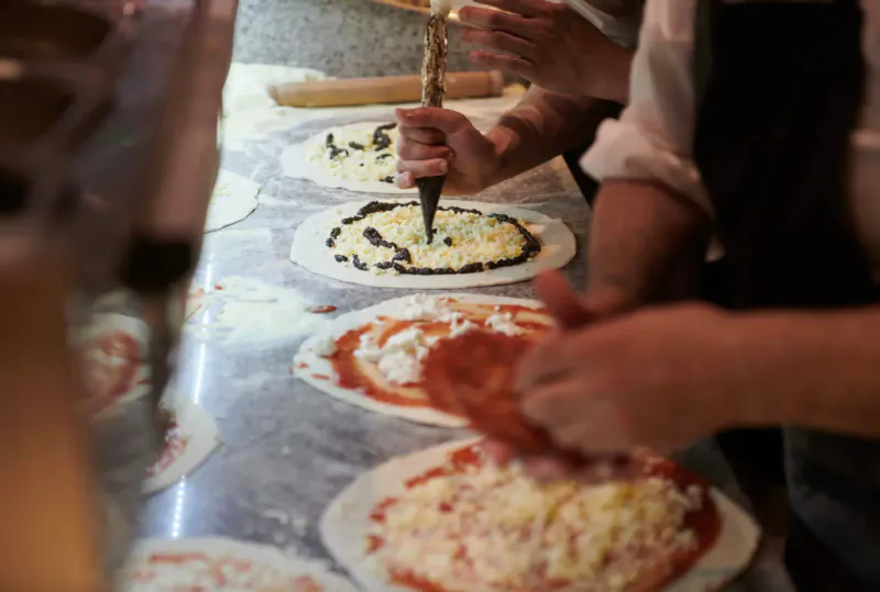 Hands topping pizzas with sauce, cheese, and veggies on a flour-dusted counter for National Pizza Day.