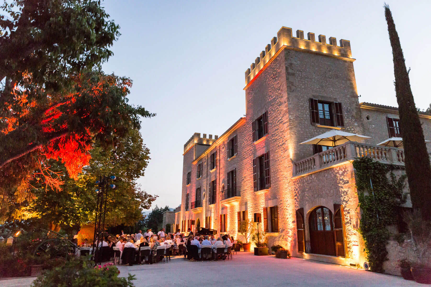 Evening wedding dinner at Castell Son Claret, luxury stone castle in Mallorca with lit towers, trees, and guests at candlelit tables.