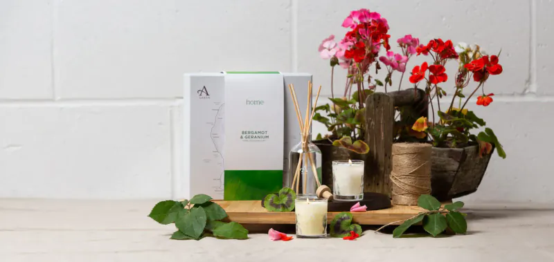 Arranged spring home fragrance display: white A'kin reed diffuser box, sticks, candle, pink geraniums in wicker pot on wood board.