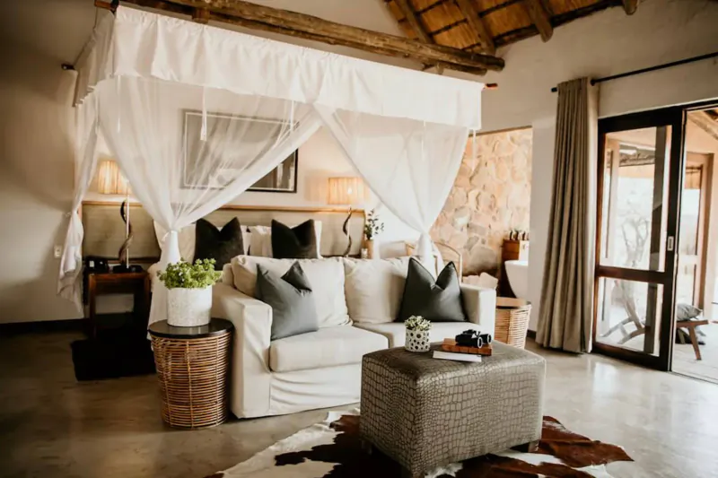 Luxurious bedroom in Nambiti Hills game reserve with white canopy bed, plush gray sofa, lamps, plants, and sliding doors to patio.