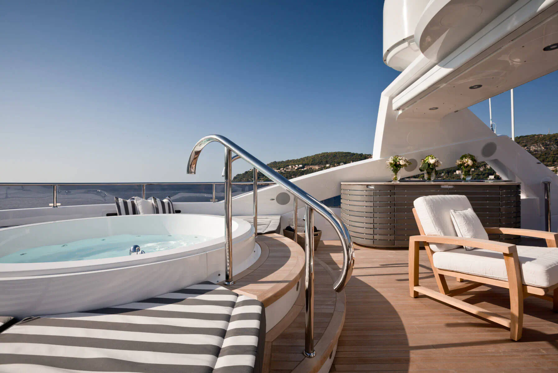 Luxury yacht deck with bubbling hot tub, chrome handrails, white chairs, flowers, and sea view under blue sky.