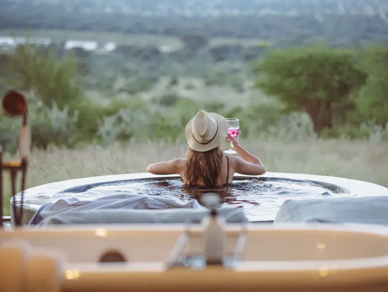 Woman in wide-brim hat sips pink cocktail in hot tub, overlooking Namibia savanna at Otjiwa.